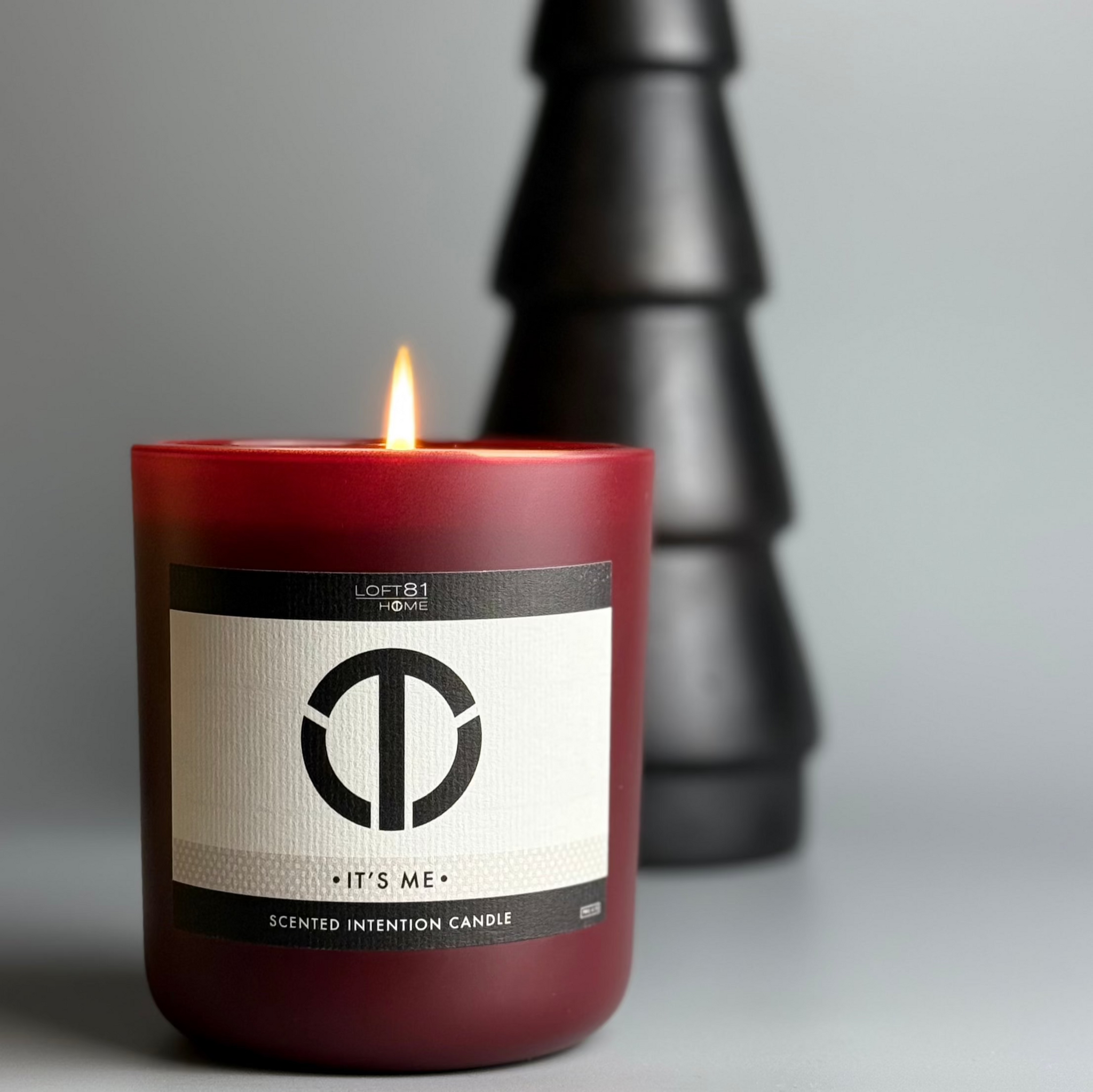 premium it’s me candle lit on a modern neutral surface beside a matte black minimalist christmas tree with warm soft lighting and sensual fragrance notes of carnal white florals decadent plum and sexy black rose creating a refined festive home ambiance